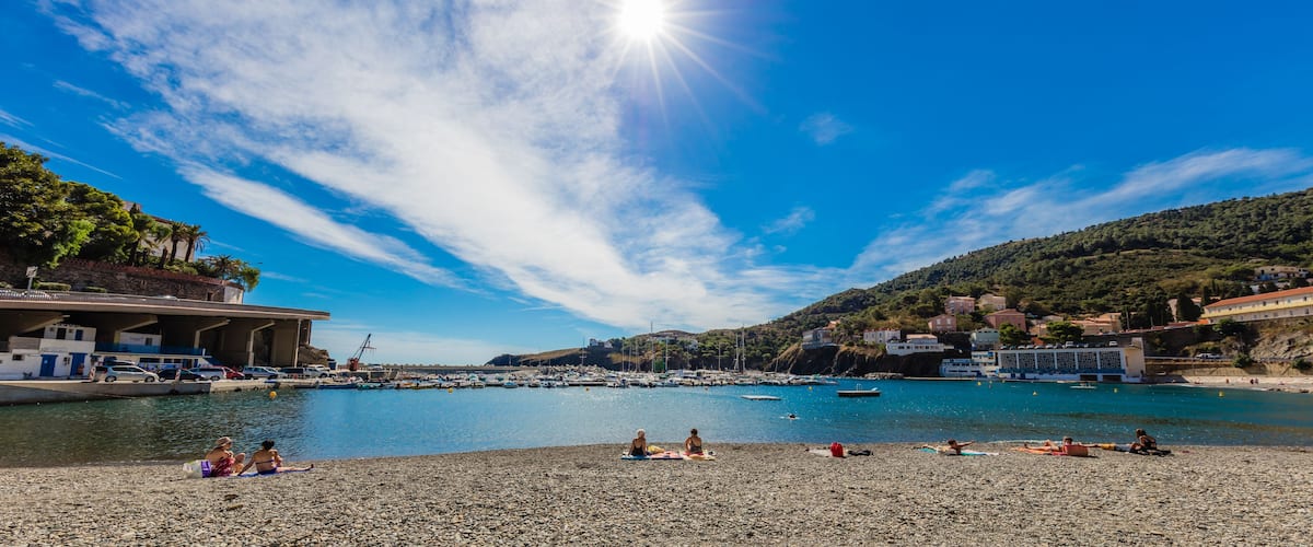 A sunny beach with pebbles, boats, and buildings in the background.
