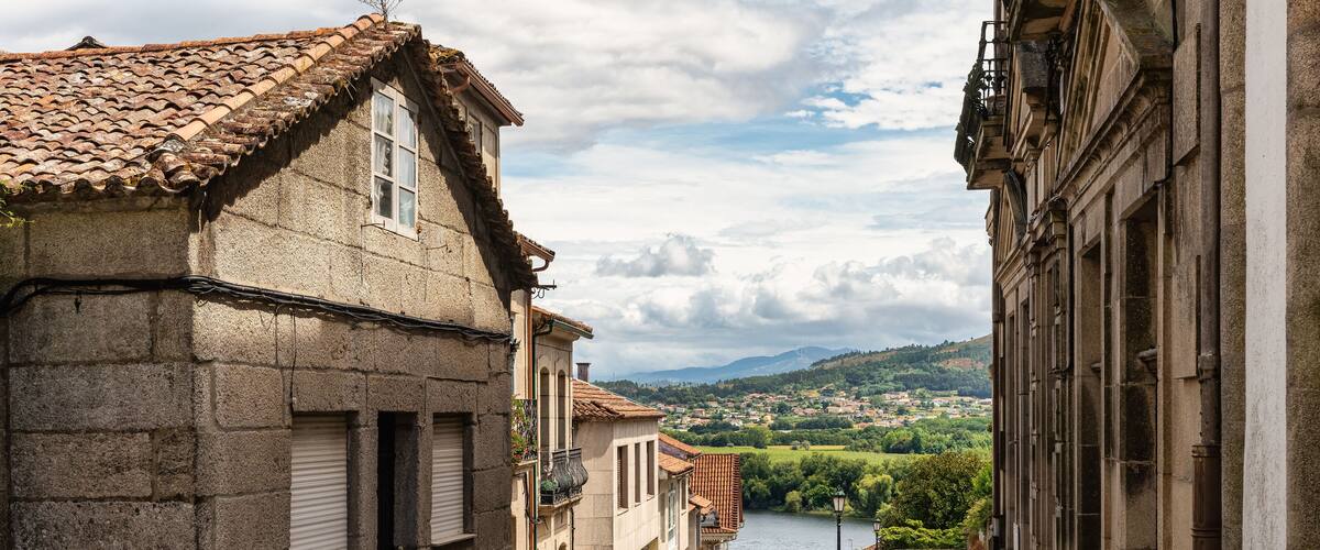 A narrow stone street flanked by historic buildings with a river and hills in the background.