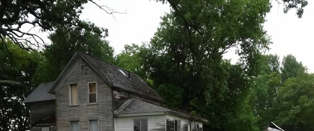 An abandoned wooden house with a dilapidated roof, surrounded by overgrown grass and a muddy path.