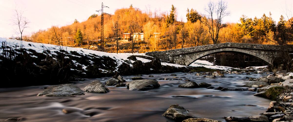 A stone bridge over a flowing river with snow-covered banks and trees in the background.