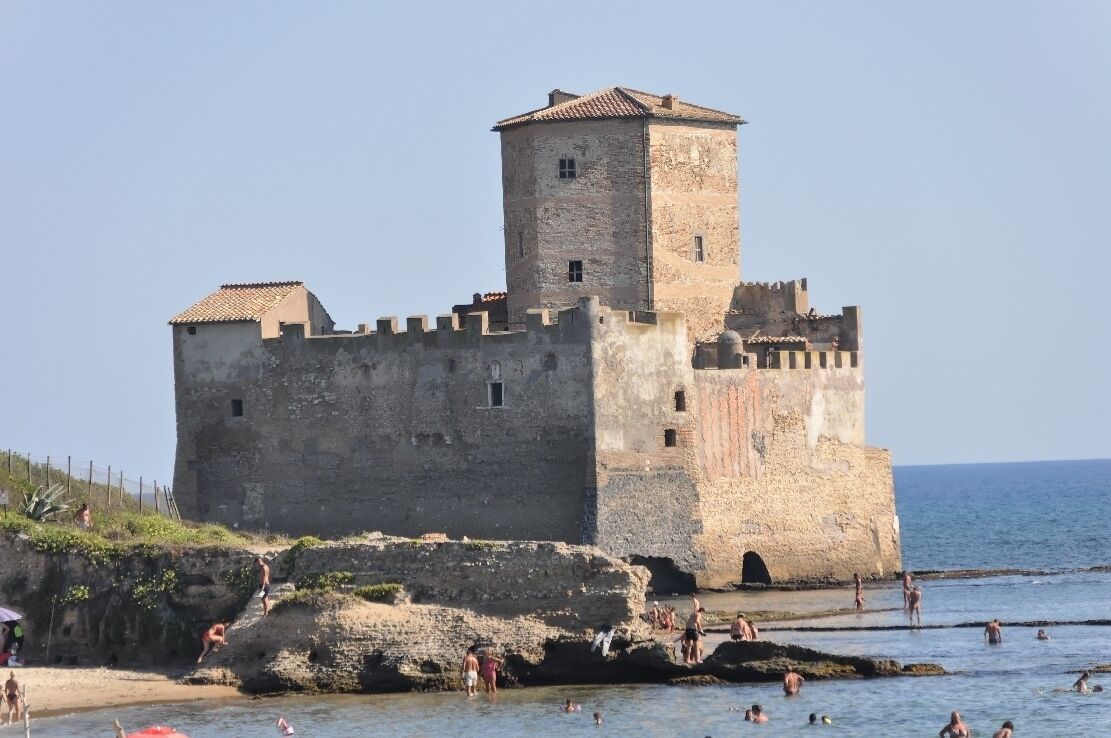 A historic stone castle by the sea with people swimming and sunbathing nearby.