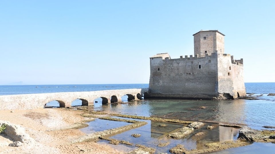 A coastal fortress with a tower, surrounded by clear waters and a sandy beach.