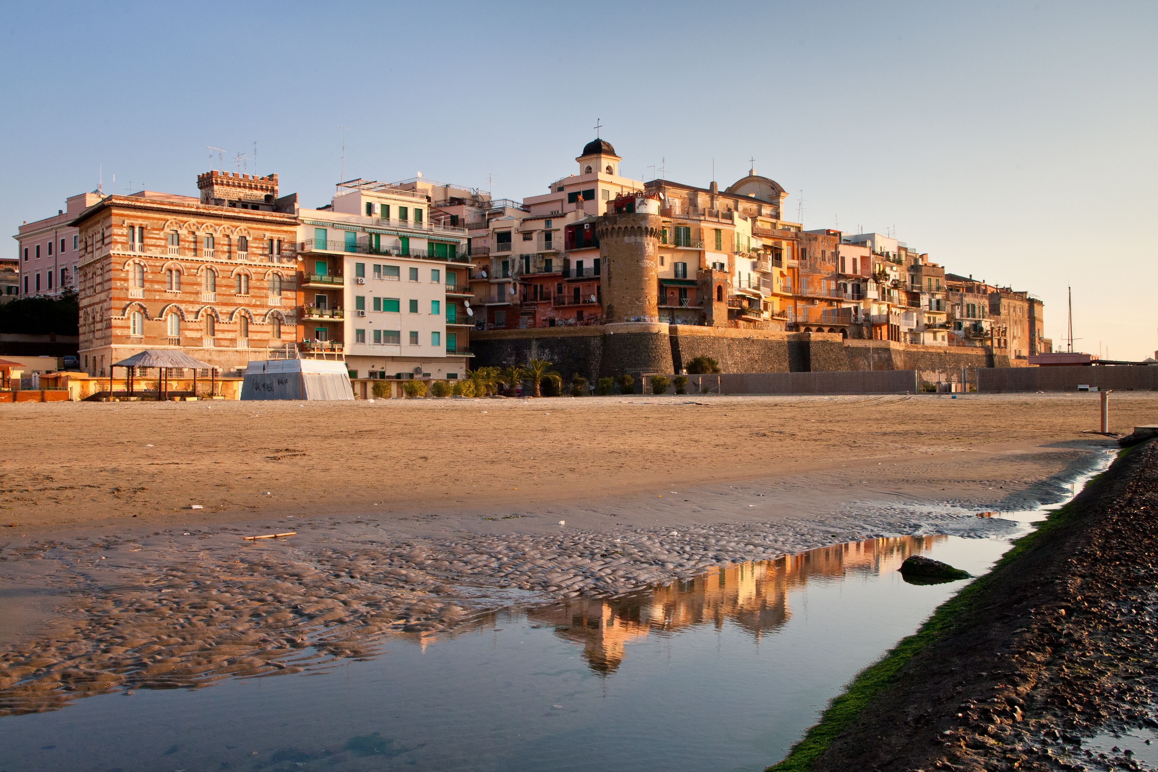 A coastal town with historic buildings, a prominent tower, and a reflective puddle on the beach.