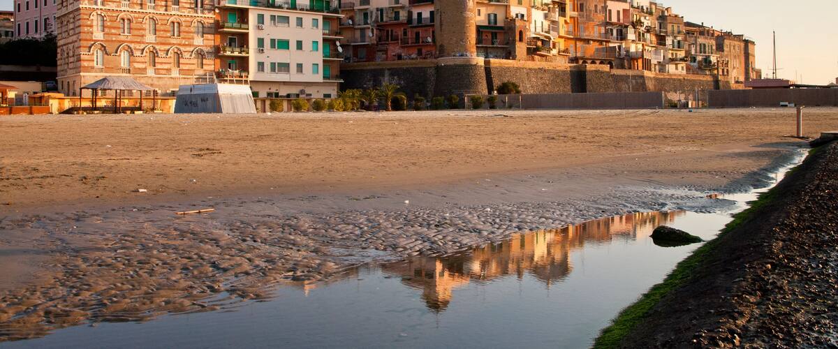 A coastal town with historic buildings, a prominent tower, and a reflective puddle on the beach.