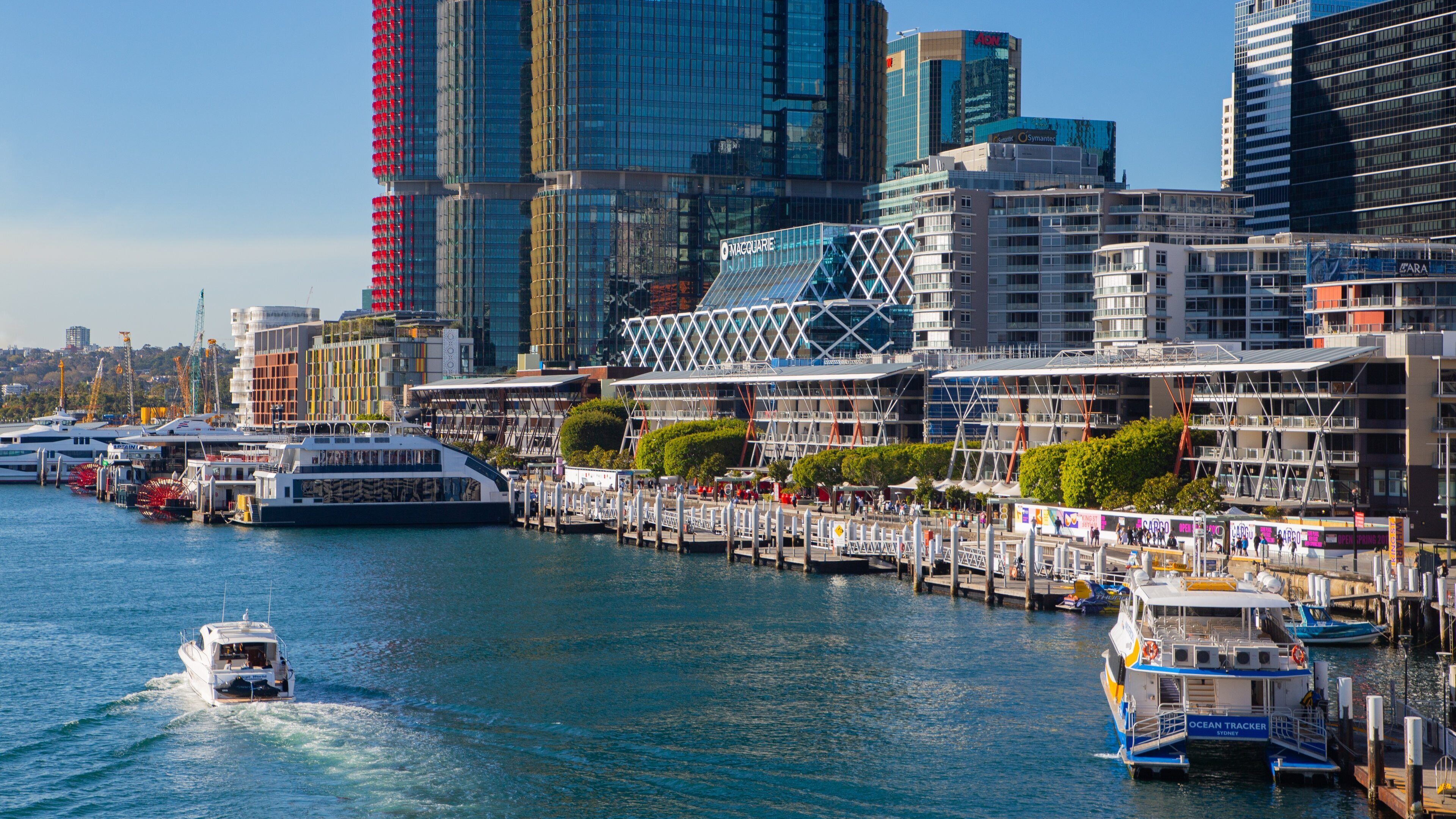 Un paysage urbain au bord de l’eau, avec des bâtiments modernes, une marina remplie de bateaux et une promenade piétonne longeant le rivage.
