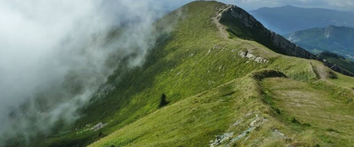 A grassy mountain ridge with a path, surrounded by clouds and distant mountains.