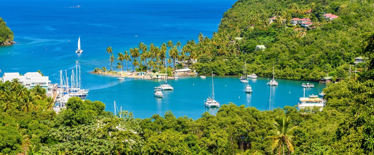 A coastal bay with numerous sailboats, a small island with buildings, and lush green hills.