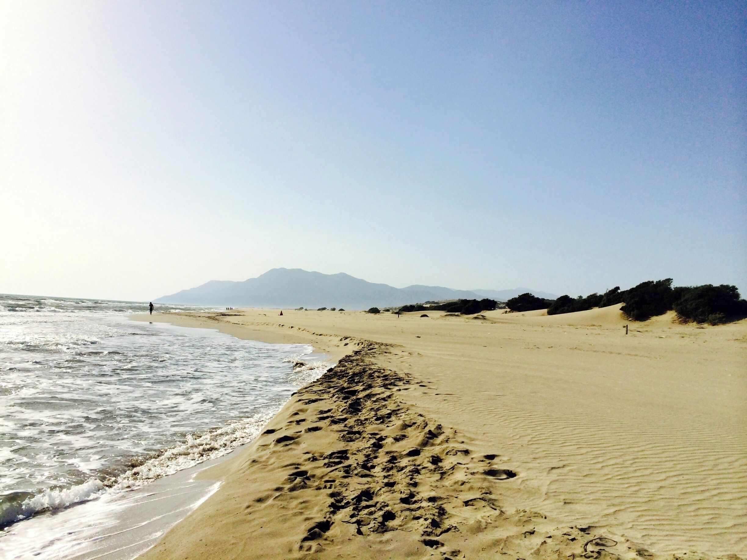 A beach with footprints leading to the water, a clear sky, and distant mountains.