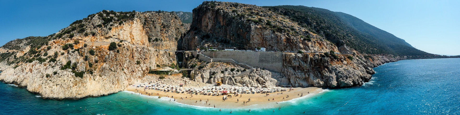A coastal landscape with a rocky cliff, a beach, and a clear blue sea.