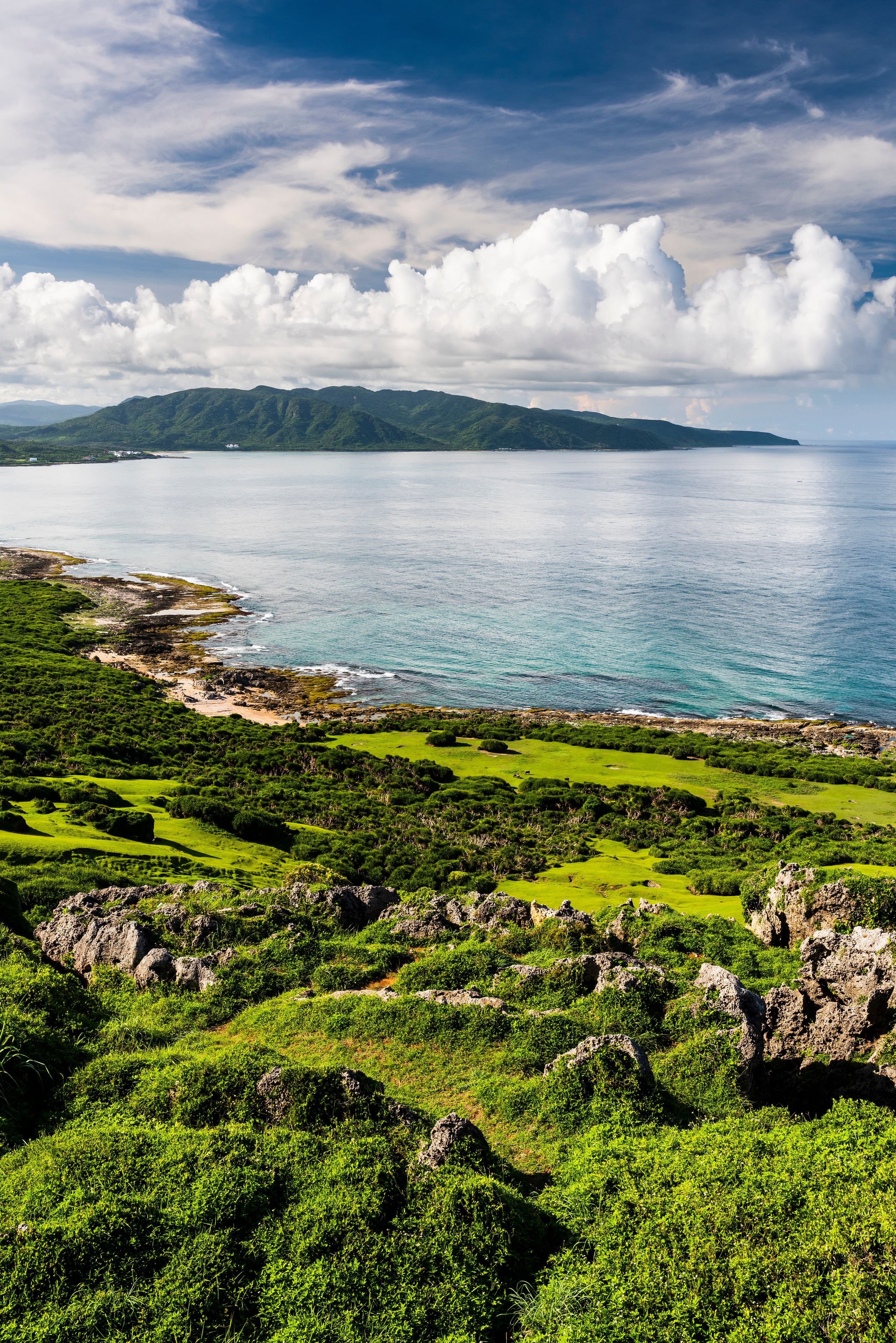 Un paisaje costero con colinas verdes, un mar azul cristalino y una cadena montañosa distante bajo un cielo parcialmente nublado.