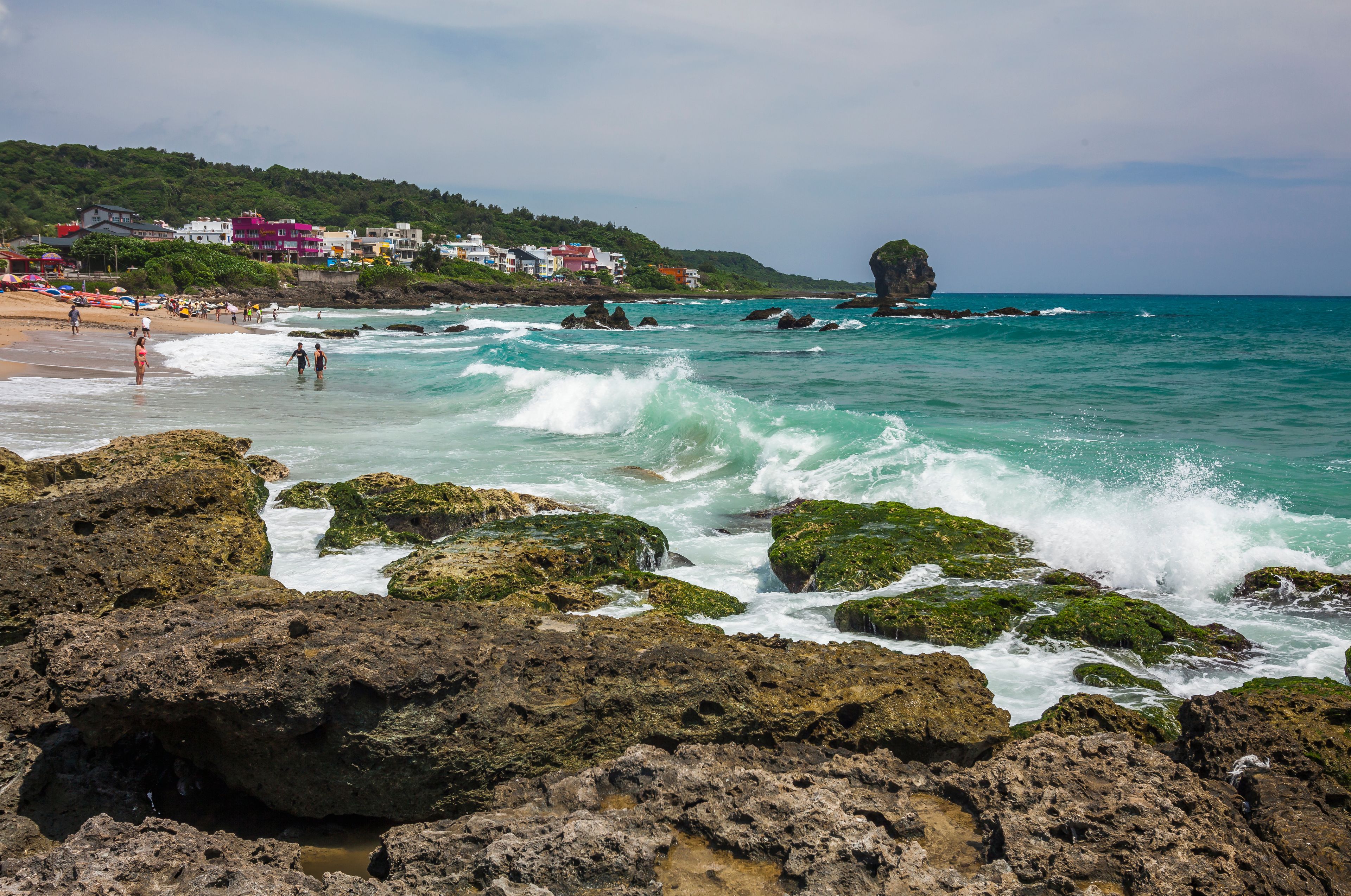 Una costa rocosa con olas rompiendo en la playa, un mar azul cristalino y edificios en una colina al fondo.
