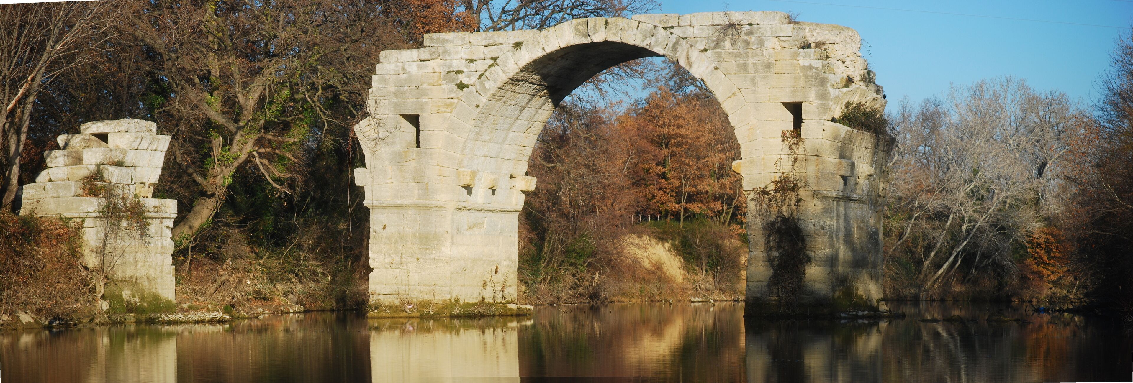 An ancient stone bridge over a calm river, surrounded by autumn trees.