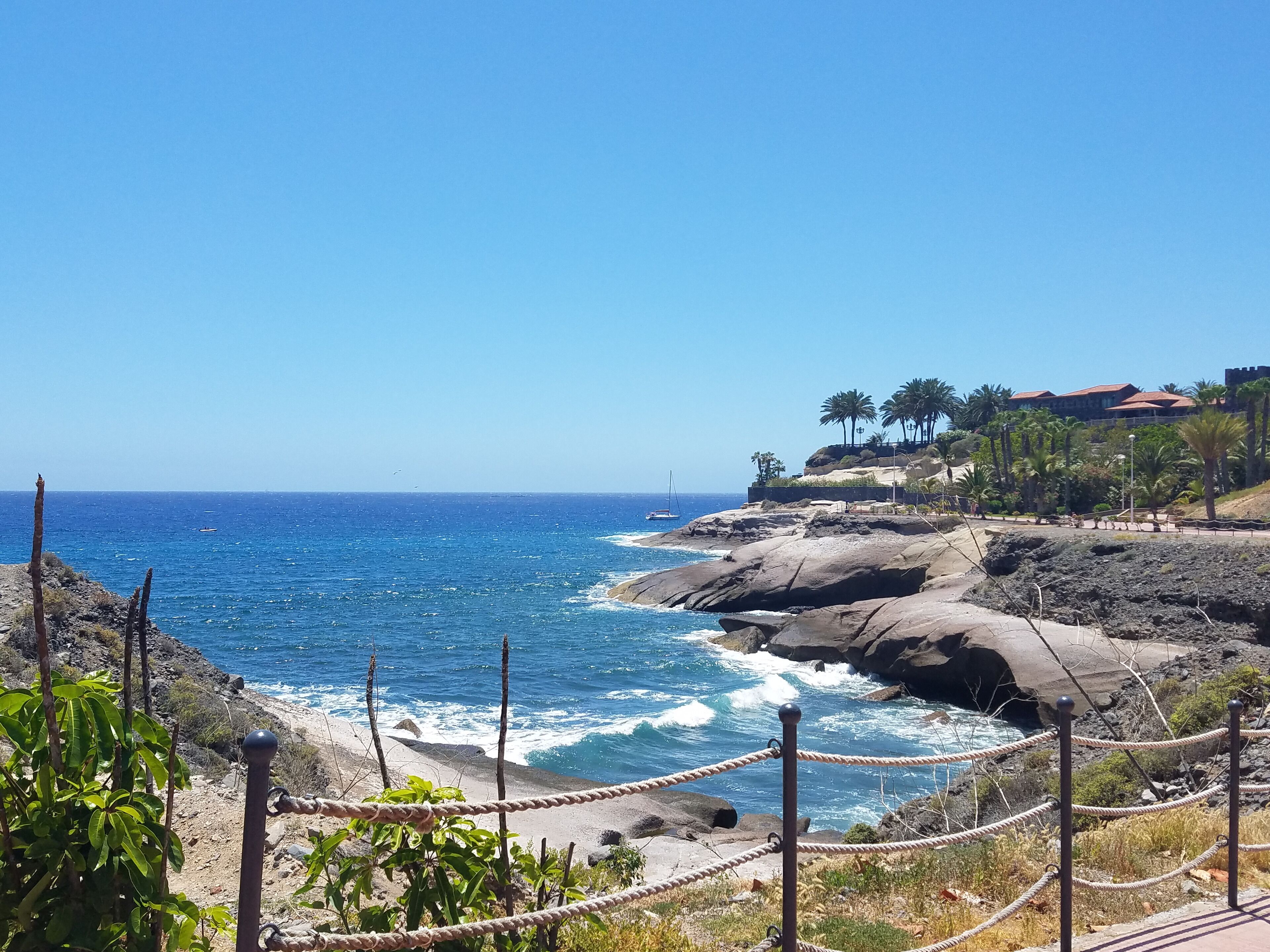 A coastal landscape with rocky cliffs, a clear blue sea, and a few sailboats.