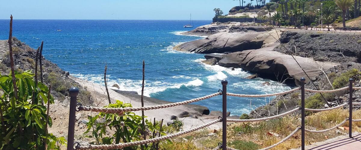A coastal landscape with rocky cliffs, a clear blue sea, and a few sailboats.