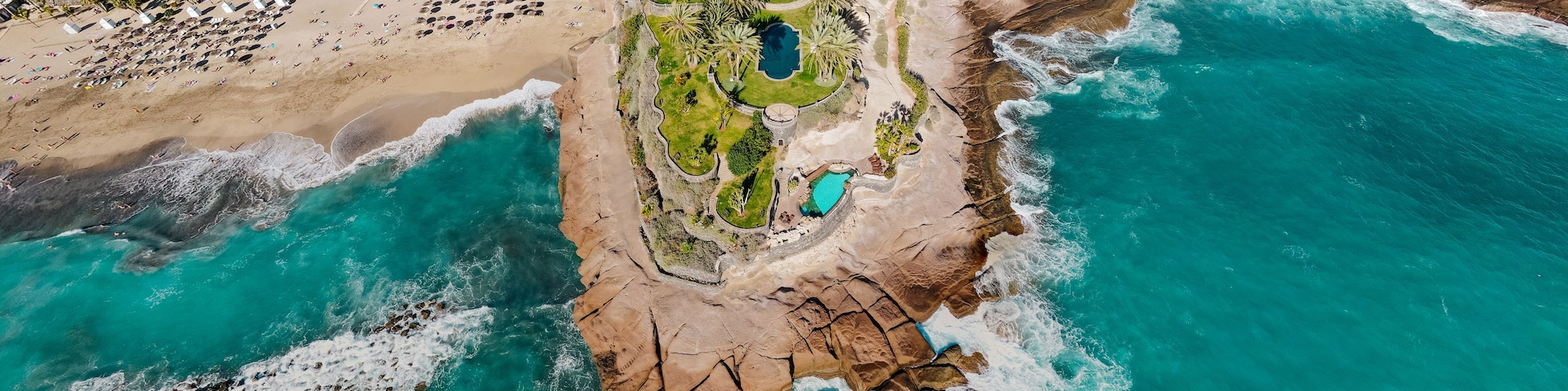 Vista aérea de un complejo turístico frente al mar, con playa, costa rocosa y una amplia zona de piscina.