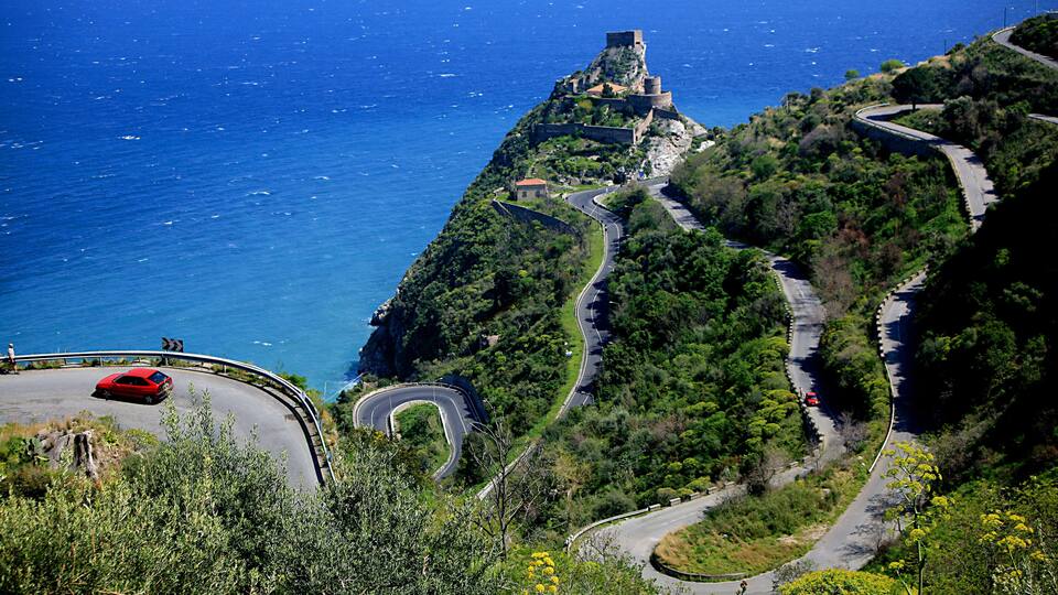 A winding mountain road with a red car, a historic castle on a hill, and the ocean in the background.