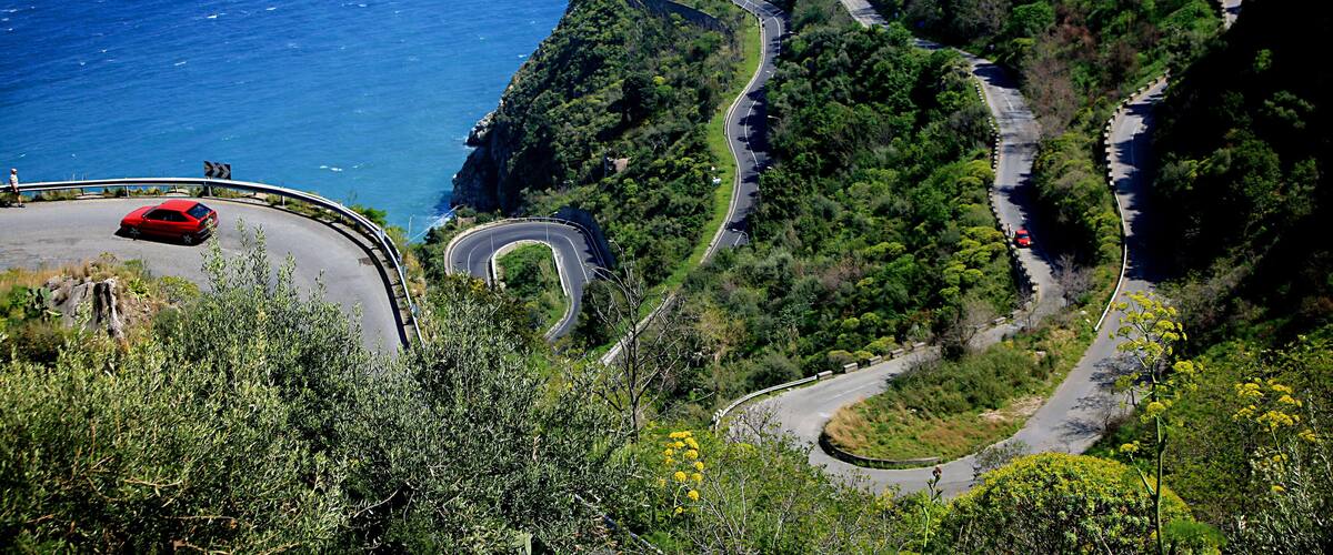 A winding mountain road with a red car, a historic castle on a hill, and the ocean in the background.