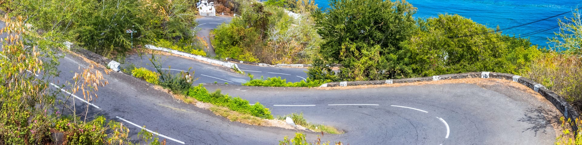 A coastal road curving through a hilly landscape with a view of the ocean and a residential area in the distance.