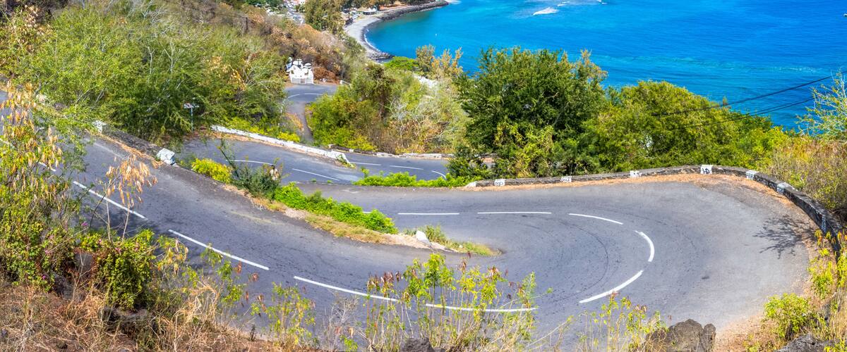 A coastal road curving through a hilly landscape with a view of the ocean and a residential area in the distance.
