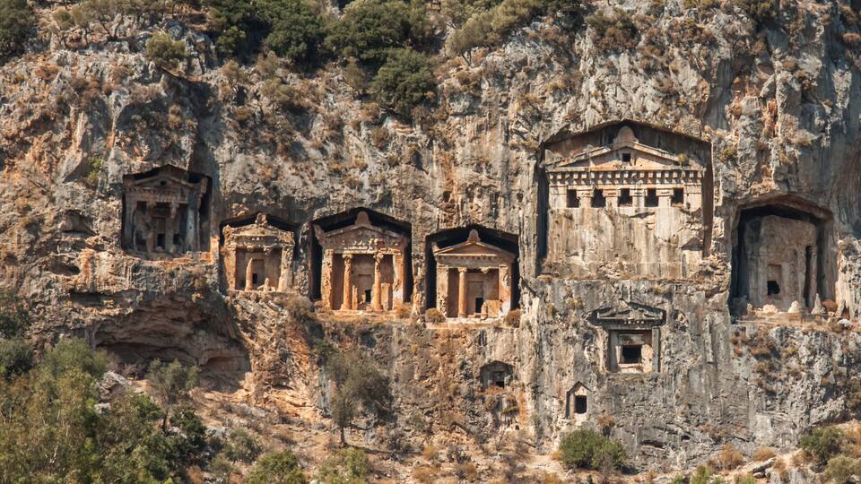 Ancient ruins carved into a cliffside, featuring multiple structures with columns and a central building with a pediment.
