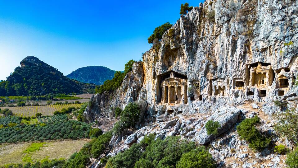 A cliff with ancient rock-cut tombs, a mountainous backdrop, and a vineyard below.