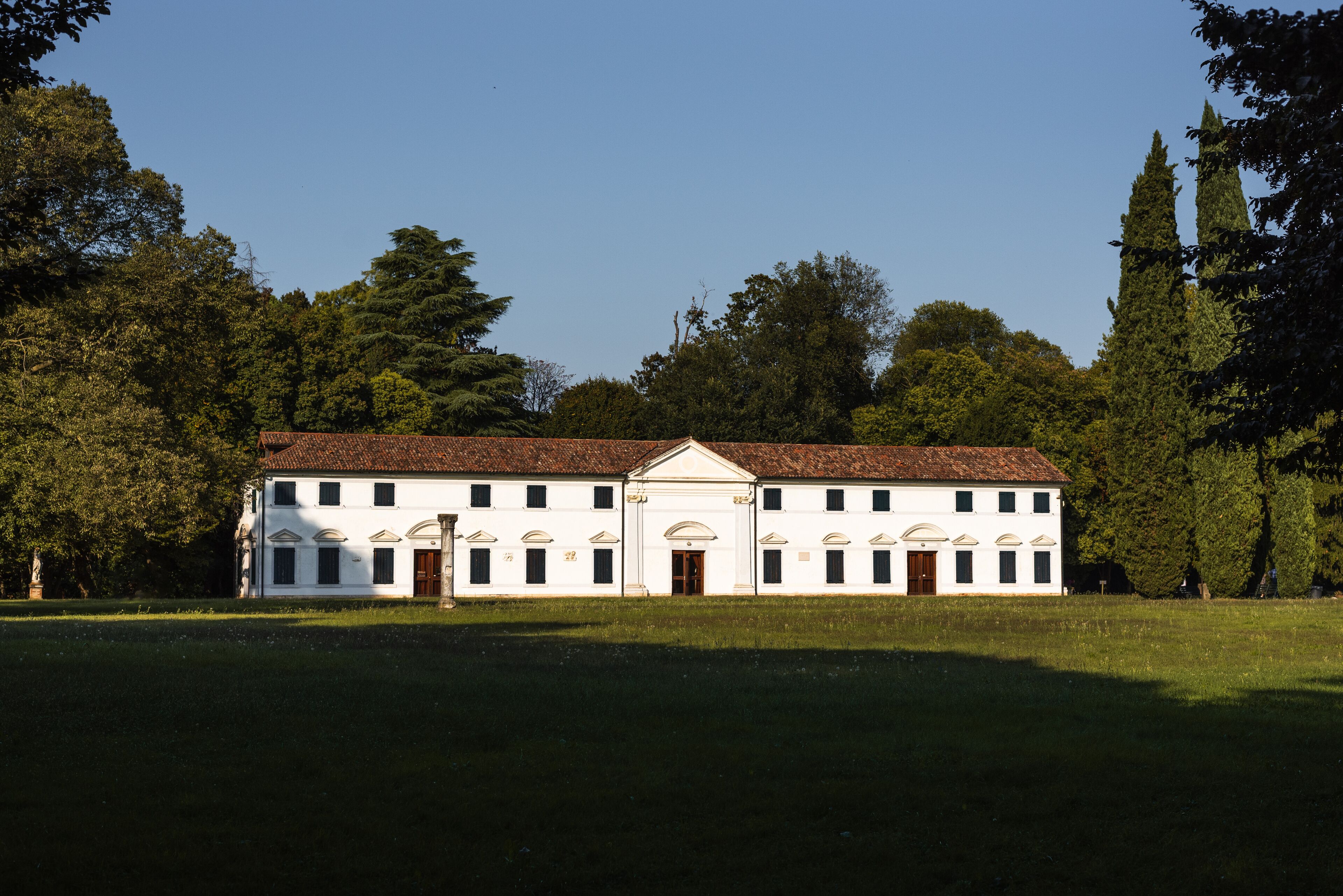 A white building with a red-tiled roof, surrounded by trees and grass.