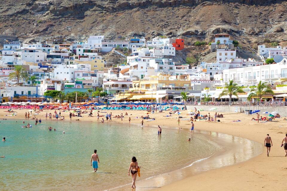 A crowded beach with people sunbathing and swimming, surrounded by colorful buildings and palm trees.