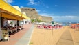 A beach with red and white sun loungers, a wooden boardwalk, and a rocky cliff in the background.