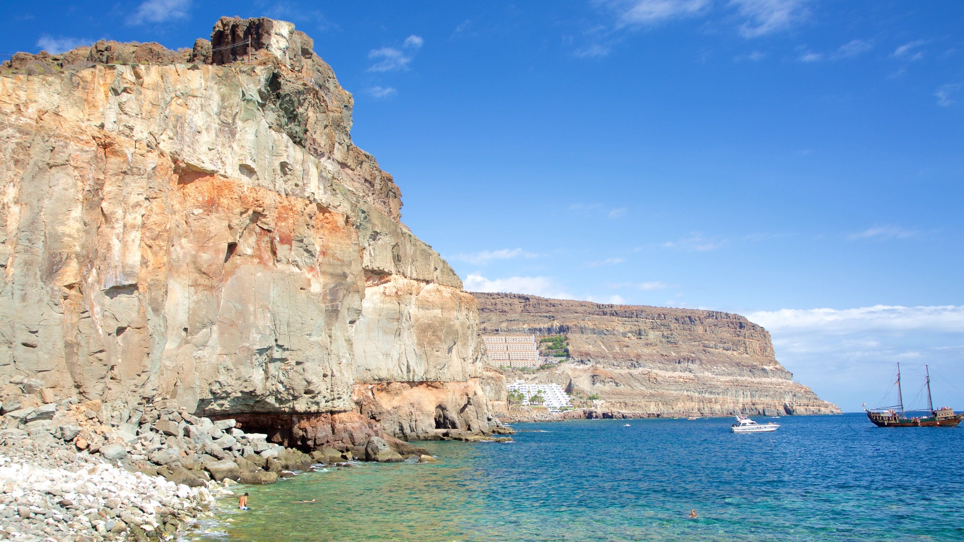 A rocky coastline with clear blue waters, a large cliff, and a few boats.
