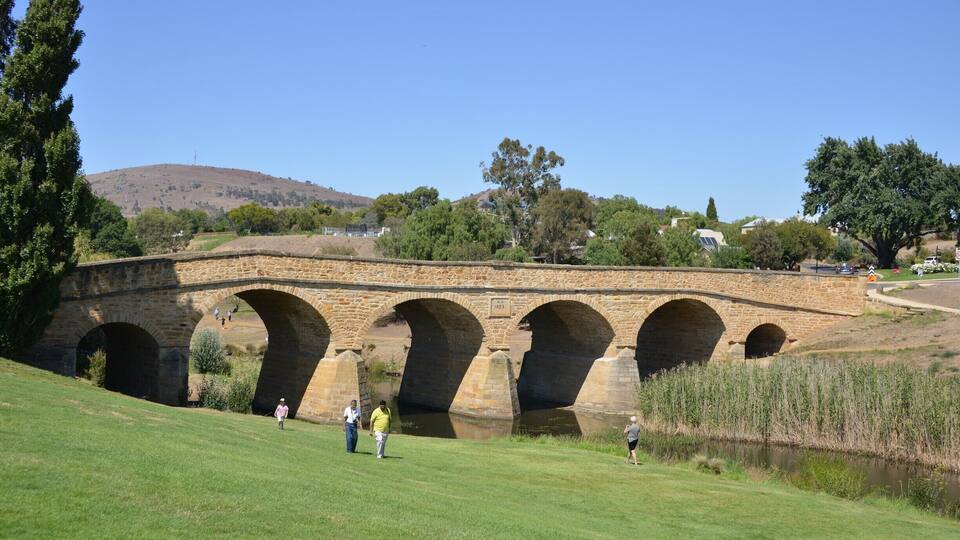 Eine Steinbrücke mit mehreren Bögen über einem Fluss, umgeben von Grünflächen und Hügeln.