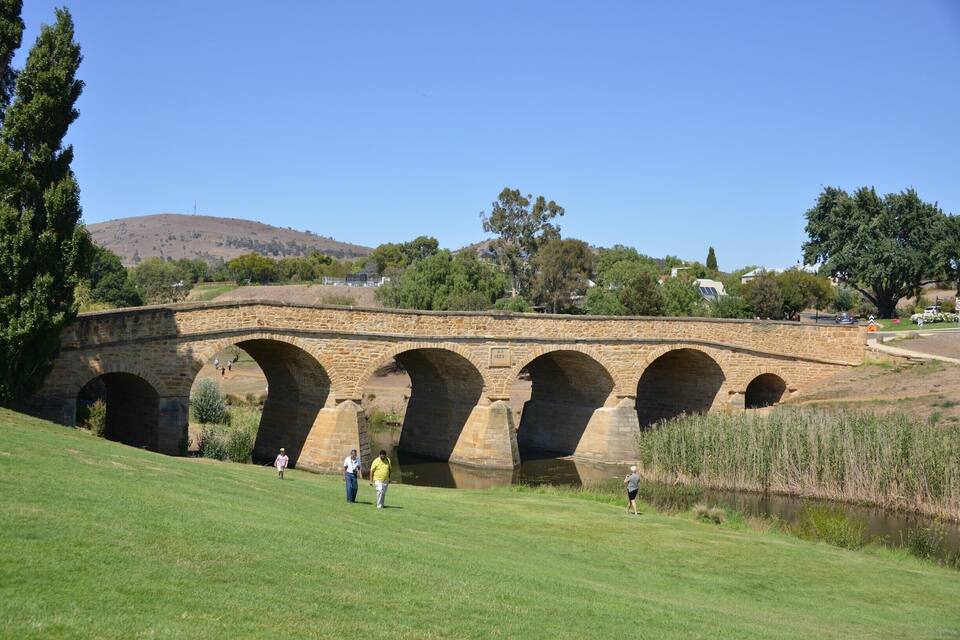 A stone bridge with multiple arches over a river, surrounded by greenery and hills.