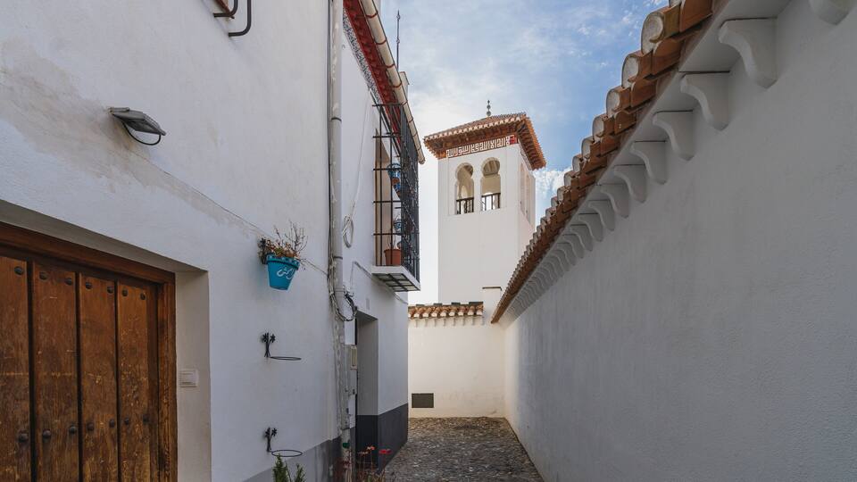 A narrow cobblestone alleyway flanked by white buildings with terracotta roofs and a small tower with arched windows.