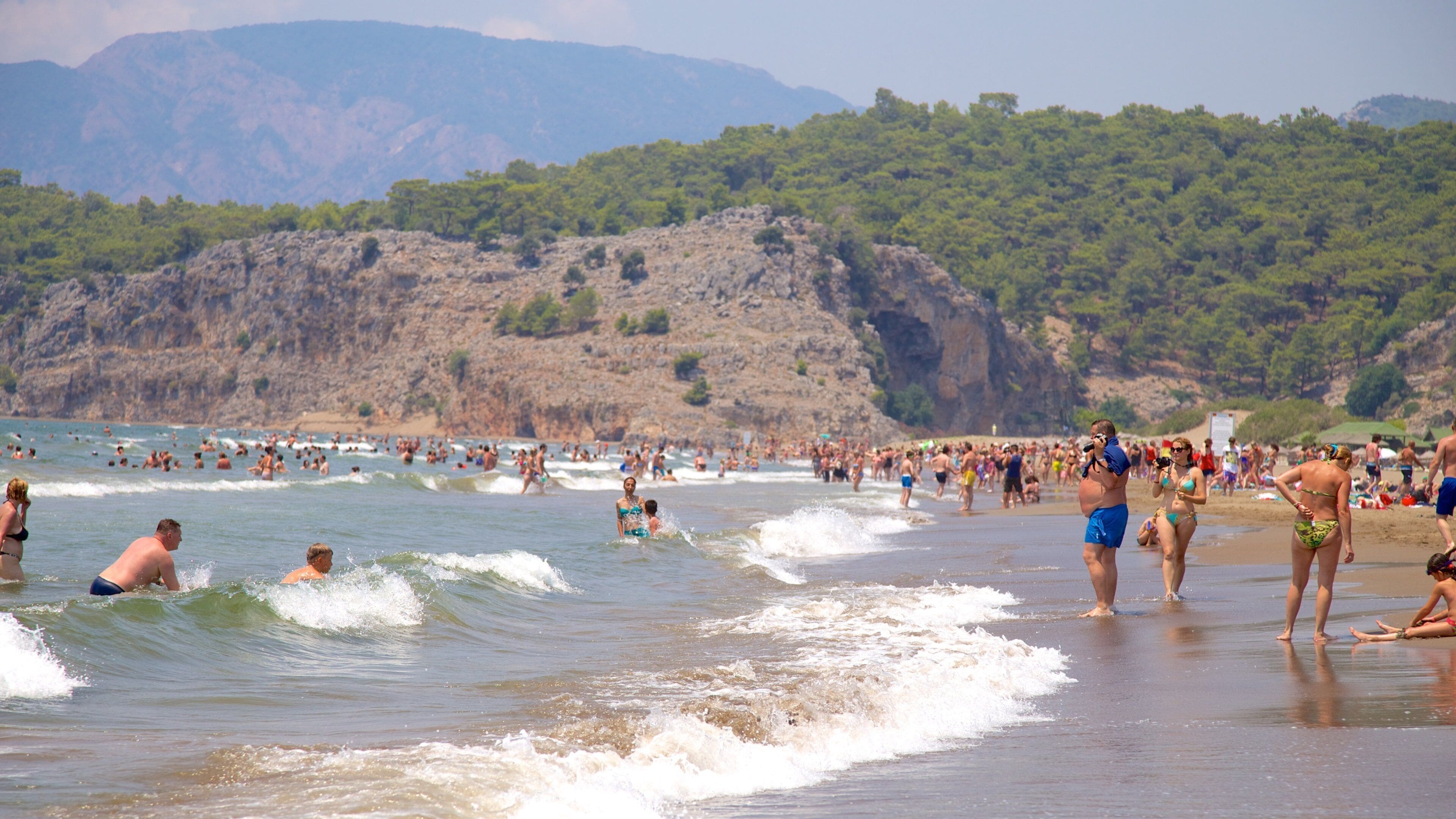 Una playa concurrida donde personas nadan y pasean por la orilla, rodeada de acantilados rocosos y abundante vegetación.