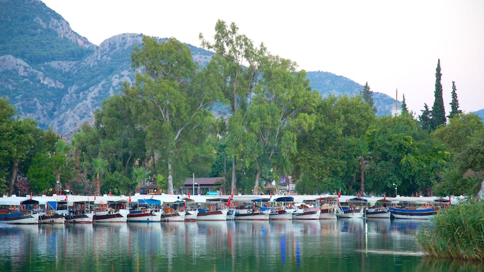 A calm lake with several boats docked along the shore, surrounded by lush green trees and mountains in the background.