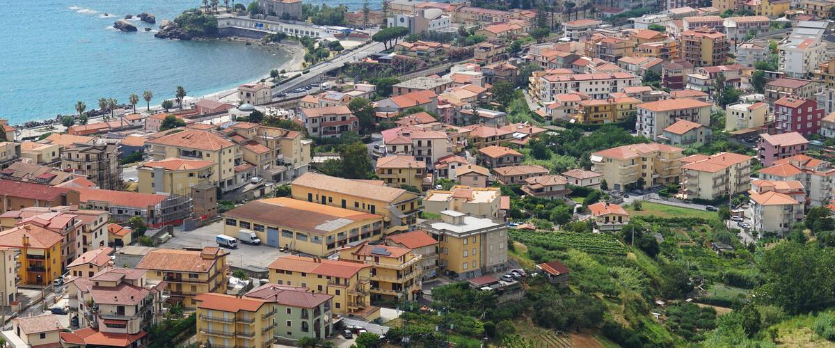 A coastal town with terracotta-roofed buildings, a marina, and a hillside vineyard.