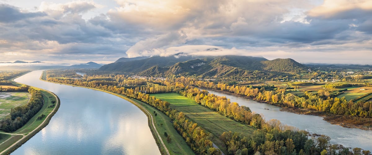 A river meandering through a valley with surrounding fields and mountains under a cloudy sky.