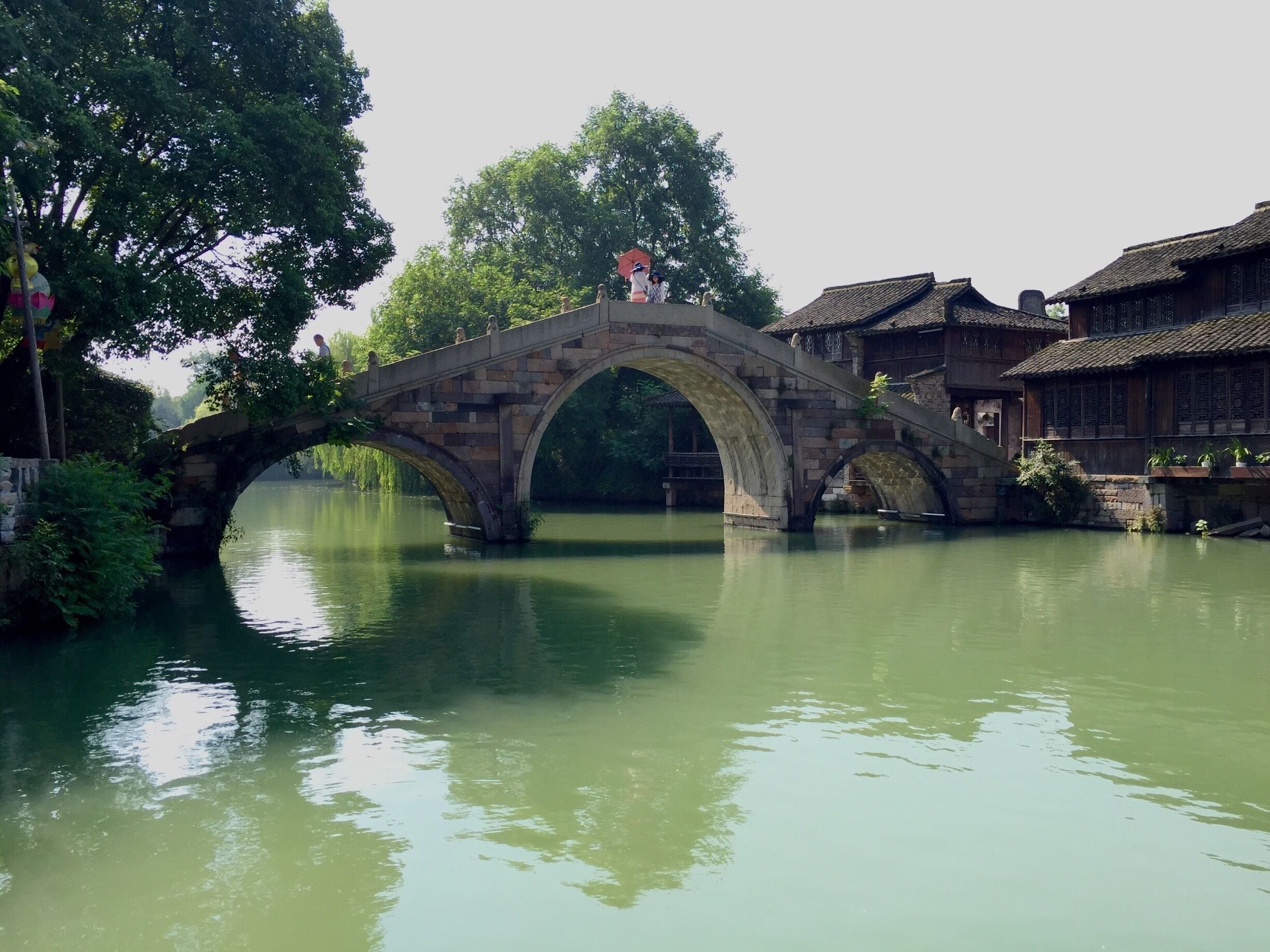 A stone bridge over a calm river, surrounded by lush greenery and traditional buildings.