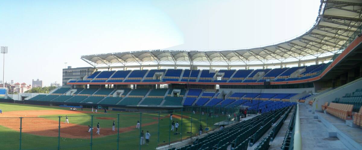 A baseball stadium with empty blue and green seats, a baseball diamond, and a large, curved roof structure.