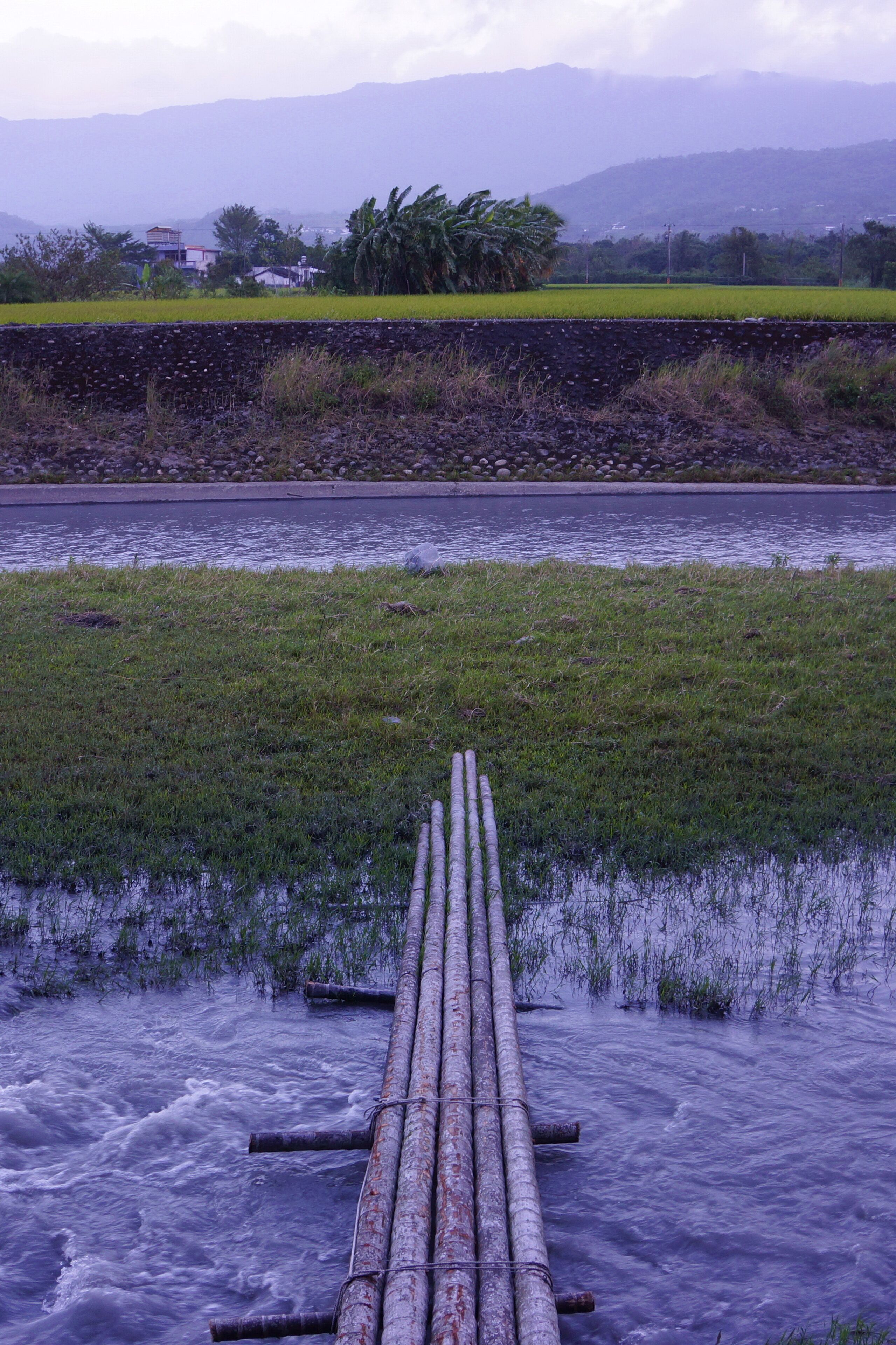 Un río con un puente de madera que lo cruza, rodeado de campos verdes y montañas al fondo.