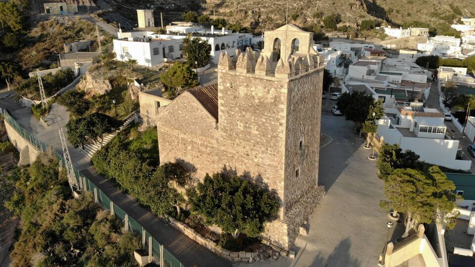 An aerial view of a historic town with a prominent stone tower, narrow streets, and surrounding greenery.