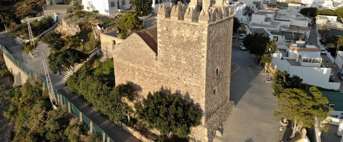 An aerial view of a historic town with a prominent stone tower, narrow streets, and surrounding greenery.