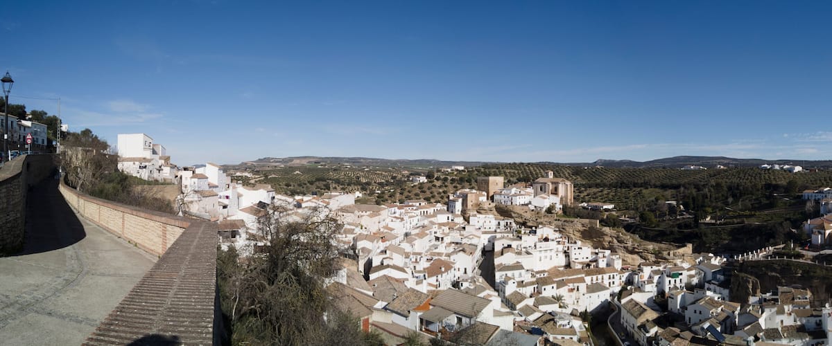 A town with white buildings on a hillside, a stone wall, and a clear blue sky.