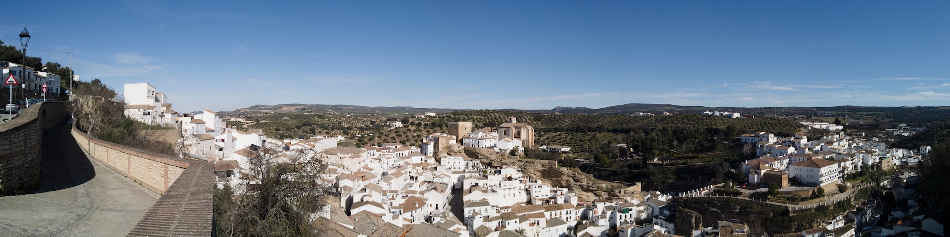 A town with white buildings on a hillside, a stone wall, and a clear blue sky.