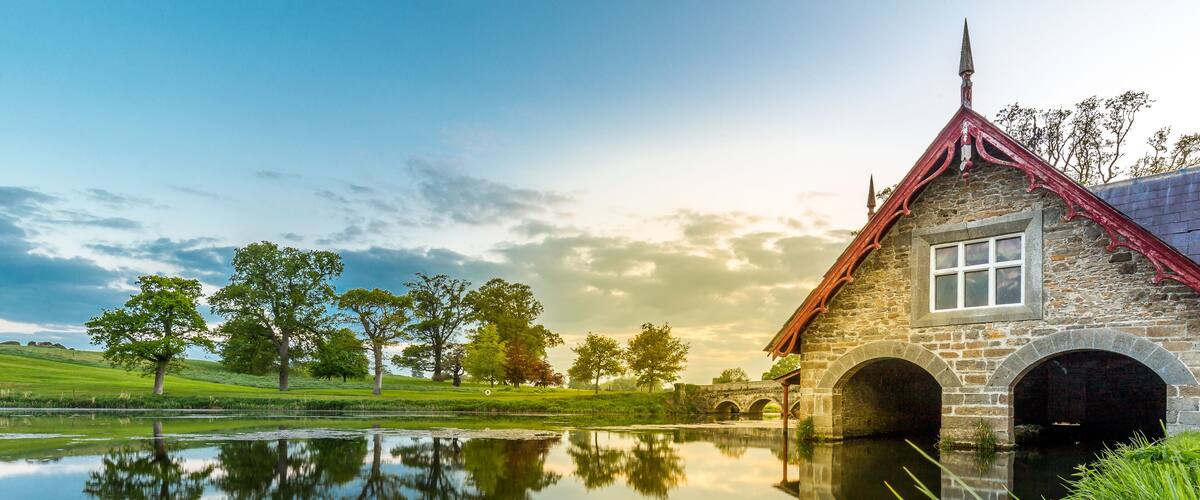 A stone building with a red roof and arched entrance, reflecting in a calm body of water, surrounded by green grass and trees.