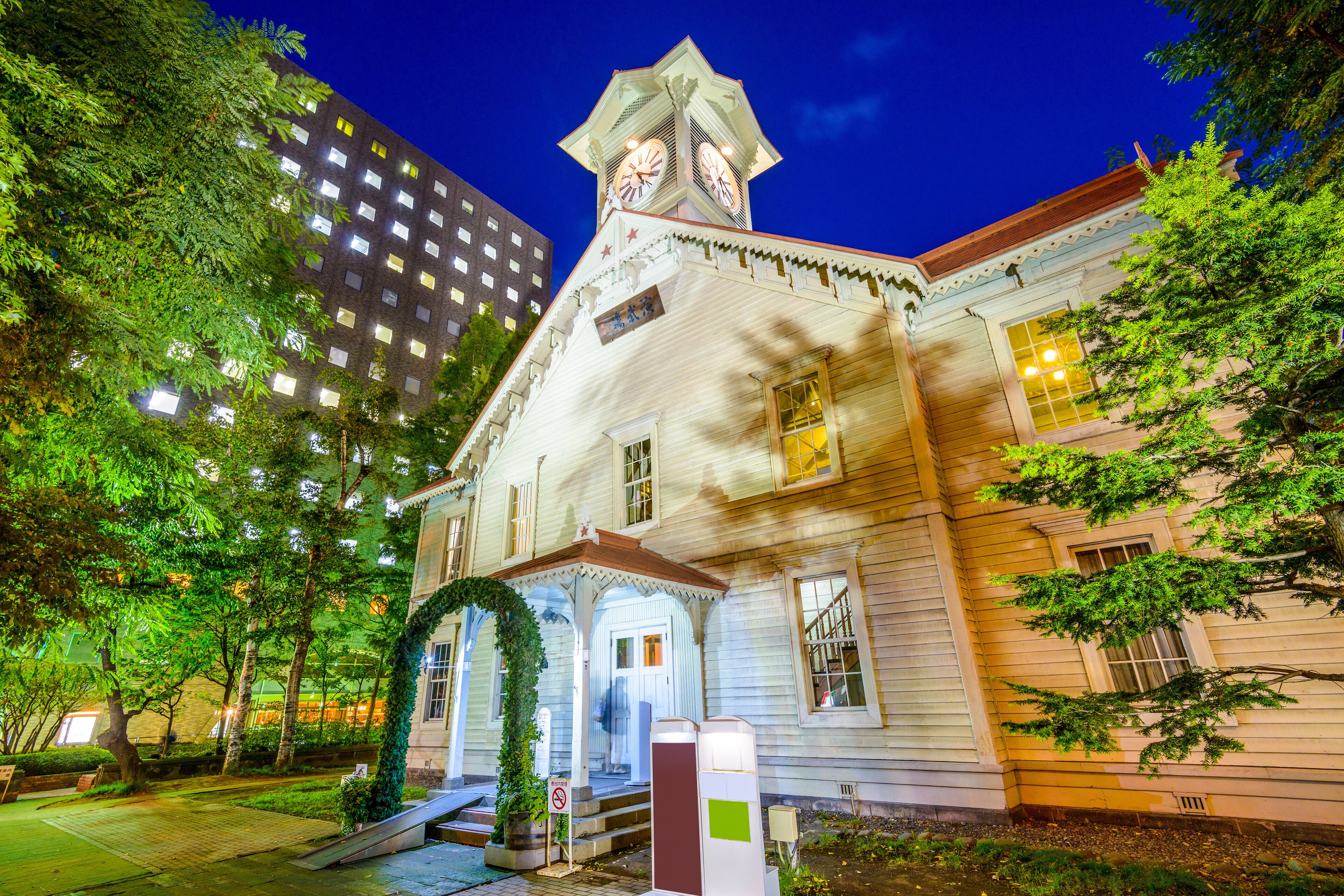 A historic building with a clock tower, surrounded by trees and a modern high-rise building.