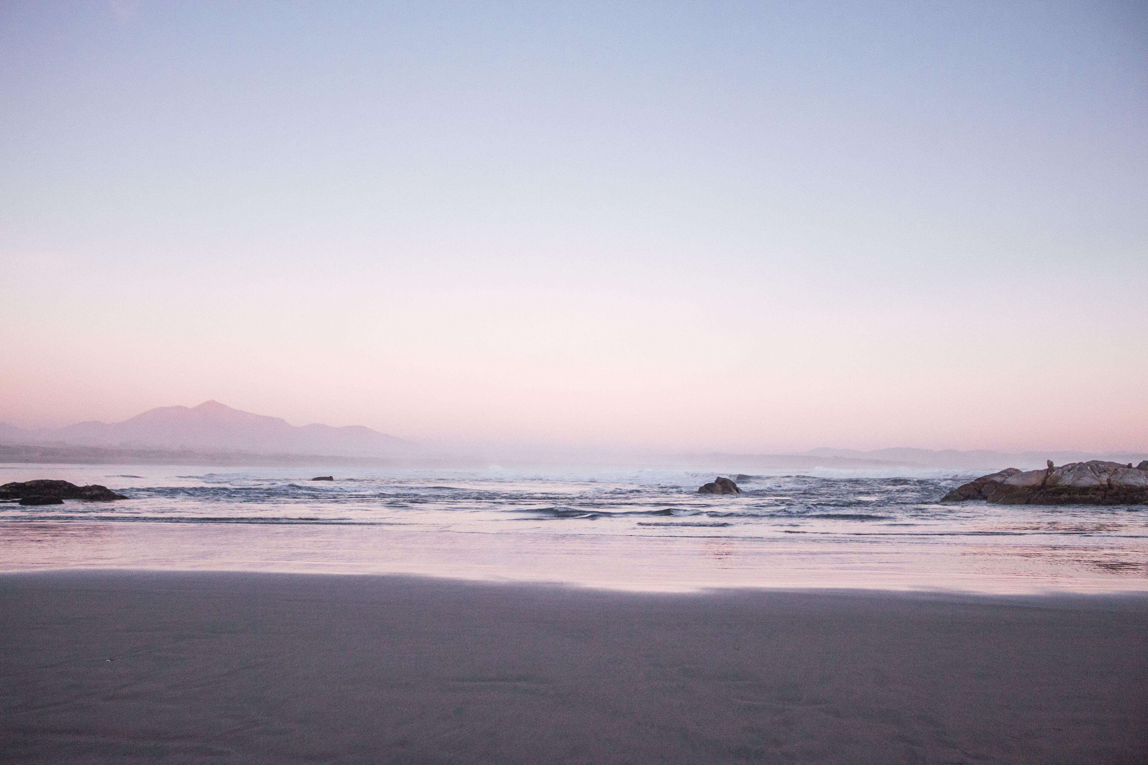 A beach with waves, rocks, and mountains in the distance.