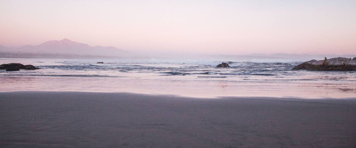 A beach with waves, rocks, and mountains in the distance.