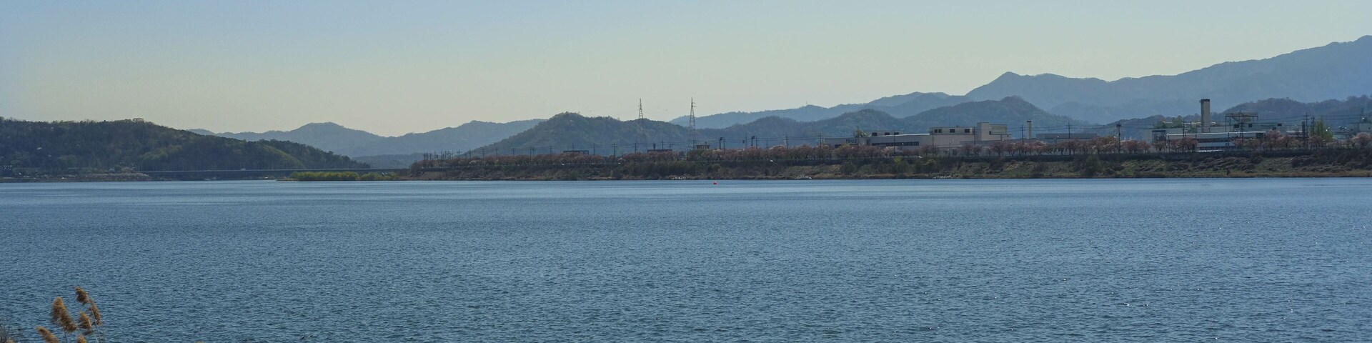 Un lago tranquilo con montañas al fondo y algunos edificios industriales cerca de la orilla.