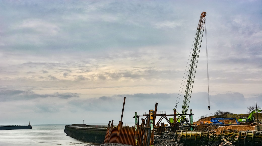 Eine Küstenbaustelle mit einem Kran, Materialhaufen und einer Wellenbrecheranlage im Hintergrund.