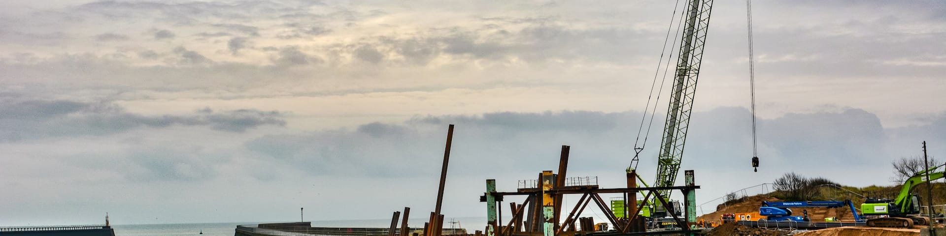 A coastal construction site with a crane, piles of materials, and a breakwater structure in the background.